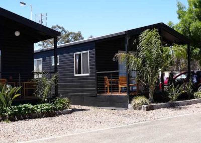 Two adjoining black cabins with small decks and outdoor seating face a gravel pathway. A red vehicle is partially visible on the right side. One of the Cabins, Dwellings, Caravans, & Glamping Tents at Stawell Grampians Gate C&C Park.