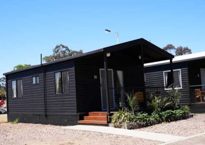 A black cabin with a gabled roof and small front deck features a mesh screen door, outdoor lighting, and surrounding garden beds with small palms and shrubs. One of the Cabins, Dwellings, Caravans, & Glamping Tents at Stawell Grampians Gate C&C Park.