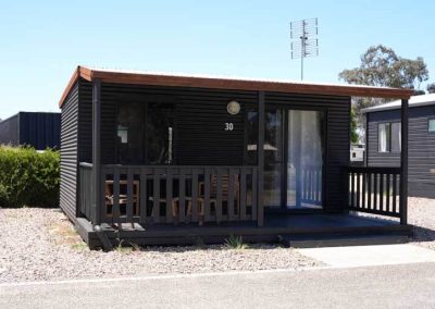 A small black cabin with horizontal cladding, a covered front deck, and sliding glass door. The number 30 is displayed near the entrance. One of the Cabins, Dwellings, Caravans, & Glamping Tents at Stawell Grampians Gate C&C Park.