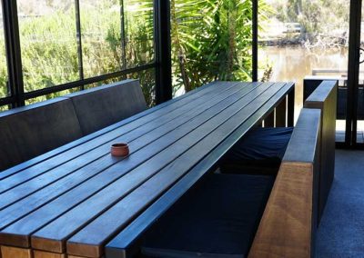 A large, dark-wood table with matching bench-style seating sits in a glass-enclosed area overlooking a pond. A small item rests at the center of the table. One of the Cabins, Dwellings, Caravans, & Glamping Tents at Stawell Grampians Gate C&C Park.