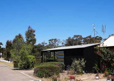 A long, low building with a light roof and black cladding, partially surrounded by dense greenery and flowering plants. An open section at the front reveals part of the interior. Located beside a quiet road under a clear sky. One of the Cabins, Dwellings, Caravans, & Glamping Tents at Stawell Grampians Gate C&C Park.