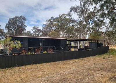 Black cabin with a raised veranda and outdoor seating, partially enclosed by a fence, surrounded by dry grass and native trees. One of the Cabins, Dwellings, Caravans, & Glamping Tents at Stawell Grampians Gate C&C Park.