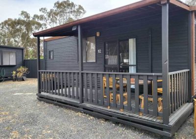 Cabin 92 with a shaded verandah and outdoor seating, nestled in a natural setting with trees and gravel path. One of the Cabins, Dwellings, Caravans, & Glamping Tents at Stawell Grampians Gate C&C Park.