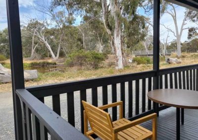 View from a cabin deck with a small round table and wooden chair, overlooking a dirt road, trees, and a calm waterway. One of the Cabins, Dwellings, Caravans, & Glamping Tents at Stawell Grampians Gate C&C Park.