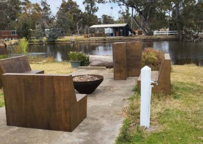 Circular fire pit surrounded by four wooden bench seats on a concrete pad beside a lake, with cabins and trees in the background at Stawell Grampians Gate C&C Park