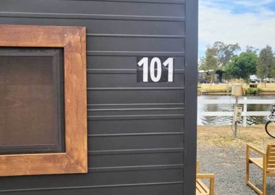 Cabin 101, identified by its bold white numbers on a modern dark exterior with a timber-framed window, sits close to the peaceful lake at Stawell Grampians Gate C&C Park. One of the Cabins, Dwellings, Caravans, & Glamping Tents at Stawell Grampians Gate C&C Park.