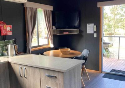 Interior of a cabin showing a kitchen counter, dining area, and mounted TV, with access to an outdoor deck at Stawell Grampians Gate C&C Park. One of the Cabins, Dwellings, Caravans, & Glamping Tents available at the park.