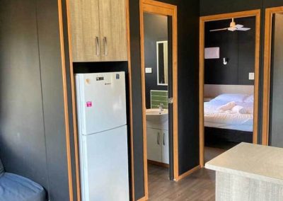 Interior view of a cabin showing a fridge, access to a bathroom, and a bedroom with a double bed at Stawell Grampians Gate C&C Park. One of the Cabins, Dwellings, Caravans, & Glamping Tents available at the park.