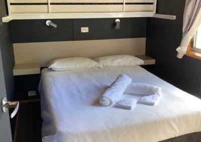 Bedroom with a double bed, overhead bunk, and built-in reading lights. Towels are placed on the bed, and a window provides natural light. One of the Cabins, Dwellings, Caravans, & Glamping Tents at Stawell Grampians Gate C&C Park.