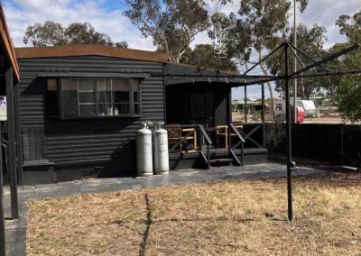 Exterior view of a black cabin with a small covered deck, outdoor seating, and dual gas cylinders. A rotary clothesline and dry grassy yard are also visible. One of the Cabins, Dwellings, Caravans, & Glamping Tents at Stawell Grampians Gate C&C Park.