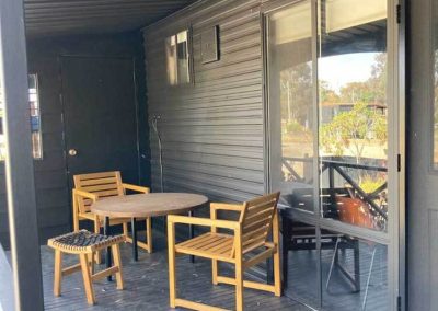 Covered deck area of a black cabin featuring a round wooden table with matching chairs and a stool. Sliding glass doors provide access to the interior. Part of the Cabins, Dwellings, Caravans, & Glamping Tents range at Stawell Grampians Gate C&C Park.