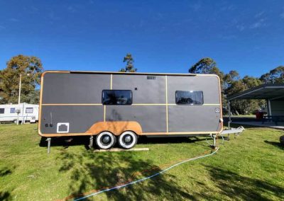 A grey retro-style caravan with timber detailing and rectangular windows, parked on grass under a clear sky at Stawell Grampians Gate C&C Park – one of the available Cabins, Dwellings, Caravans, & Glamping Tents.