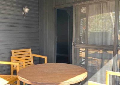 Covered outdoor deck area of a cabin with a round wooden table and wooden chairs, providing a shaded space for sitting outside. One of the Cabins, Dwellings, Caravans, & Glamping Tents options at Stawell Grampians Gate C&C Park.