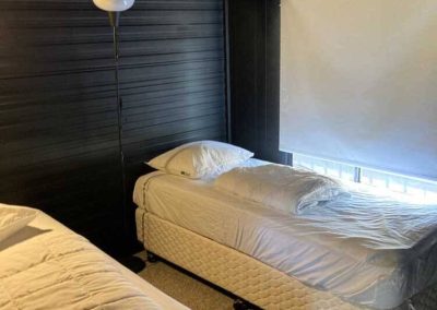 Interior bedroom of a cabin showing two single beds with white bedding, a floor lamp, and a window with a roller blind. Part of the Cabins, Dwellings, Caravans, & Glamping Tents range at Stawell Grampians Gate C&C Park.