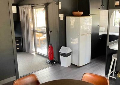 Interior view of Cabin 88 at Stawell Grampians Gate C&C Park showing a modern kitchen area with white refrigerator, red vacuum cleaner, small dining table with leather-look chairs, and a glimpse of the outdoor deck. One of the Cabins, Dwellings, Caravans, & Glamping Tents at Stawell Grampians Gate C&C Park.