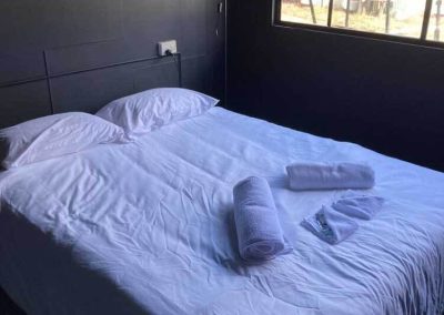 Bedroom inside Cabin 88 at Stawell Grampians Gate C&C Park, showing a double bed with crisp white linen and rolled towels, a tall floor lamp, and a large window with views of the outdoors. One of the Cabins, Dwellings, Caravans, & Glamping Tents at Stawell Grampians Gate C&C Park.