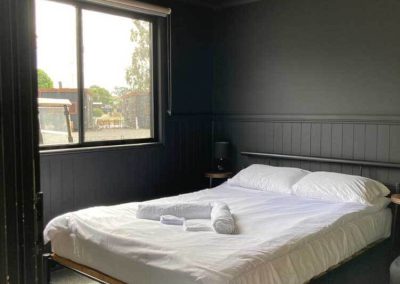 Interior of Cabin 116 at Stawell Grampians Gate C&C Park, featuring a double bed with white linen, fresh towels, a bedside table with lamp, and a large window allowing natural light into the room. One of the Cabins, Dwellings, Caravans, & Glamping Tents at Stawell Grampians Gate C&C Park.
