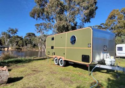 A green retro-style caravan with rounded corners, circular window, and timber trim, parked near the water at Stawell Grampians Gate C&C Park – one of the available Cabins, Dwellings, Caravans, & Glamping Tents.