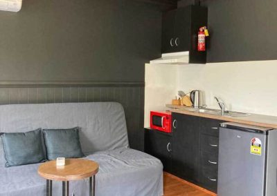 Kitchenette and lounge area inside Cabin 116 at Stawell Grampians Gate C&C Park, showing a compact kitchen with a sink, microwave, mini fridge, kettle, and overhead cabinetry, adjacent to a grey sofa with throw pillows and a small round table. One of the Cabins, Dwellings, Caravans, & Glamping Tents at Stawell Grampians Gate C&C Park.