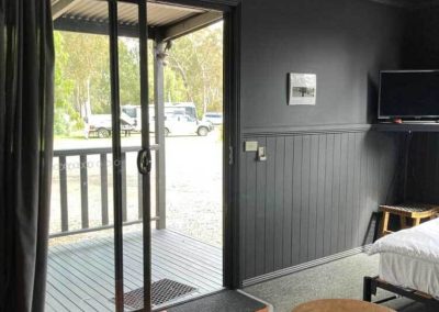 View from inside Cabin 116 at Stawell Grampians Gate C&C Park, looking out through glass sliding doors onto a private deck with timber railing, overlooking the campground and vehicles parked nearby. A TV, chair, and side table with a remote are visible in the foreground. One of the Cabins, Dwellings, Caravans, & Glamping Tents at Stawell Grampians Gate C&C Park.