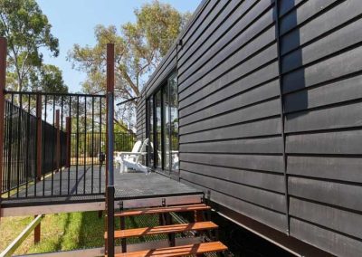 Black timber-clad exterior of a cabin with a raised deck, accessed by timber steps and featuring white outdoor seating. One of the options at Stawell Grampians Gate C&C Park – Cabins, Dwellings, Caravans, & Glamping Tents.