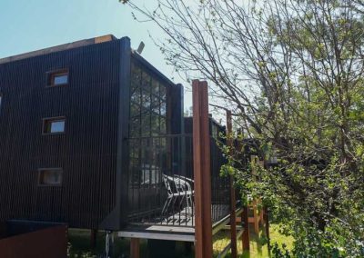 Exterior of a modern dark-toned cabin with a raised balcony and large vertical windows, surrounded by trees. One of the options at Stawell Grampians Gate C&C Park – Cabins, Dwellings, Caravans, & Glamping Tents.