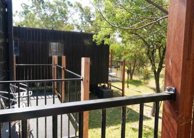 Raised deck area with metal railings and outdoor seating, overlooking another modern dark-toned cabin set among trees. One of the options at Stawell Grampians Gate C&C Park – Cabins, Dwellings, Caravans, & Glamping Tents.