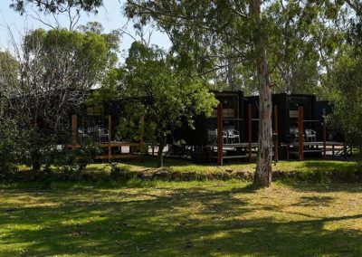 A row of modern black cabins with large glass fronts, partially hidden behind trees and greenery, viewed from across a grassy area. One of the options at Stawell Grampians Gate C&C Park – Cabins, Dwellings, Caravans, & Glamping Tents.