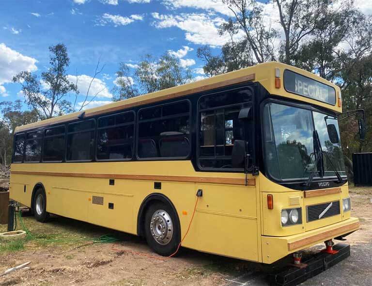 Interior view of a converted bus with two sets of wooden bunk beds on one side and a red-seated dining booth on the opposite side. A kitchenette with a red fridge and stainless steel sink is positioned beside the seating area. One of the Cabins, Dwellings, Caravans, & Glamping Tents at Stawell Grampians Gate C&C Park.