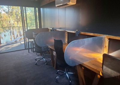 Private workstations at Stawell Grampians Gate C&C Park with office chairs in a modern room, featuring individual wooden desks separated by stainless steel dividers and a view of trees through glass doors.