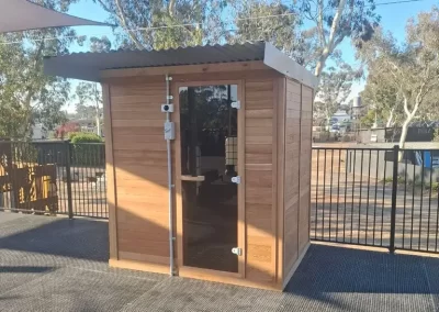 A compact wooden sauna with a glass door on a rooftop deck, surrounded by a black fence and shaded by overhead canopies.