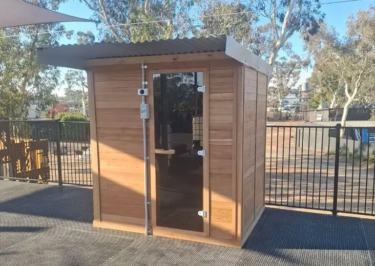 A compact wooden sauna with a glass door on a rooftop deck, surrounded by a black fence and shaded by overhead canopies.