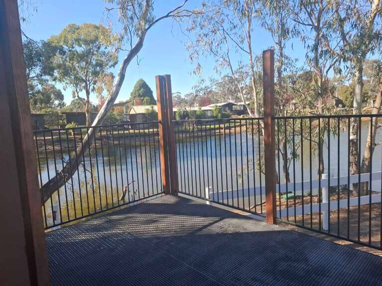 View from a balcony looking out over a small lake, with a railing in the foreground and trees, power lines, and RVs in the distance. One of the options at Stawell Grampians Gate C&C Park – Cabins, Dwellings, Caravans, & Glamping Tents.