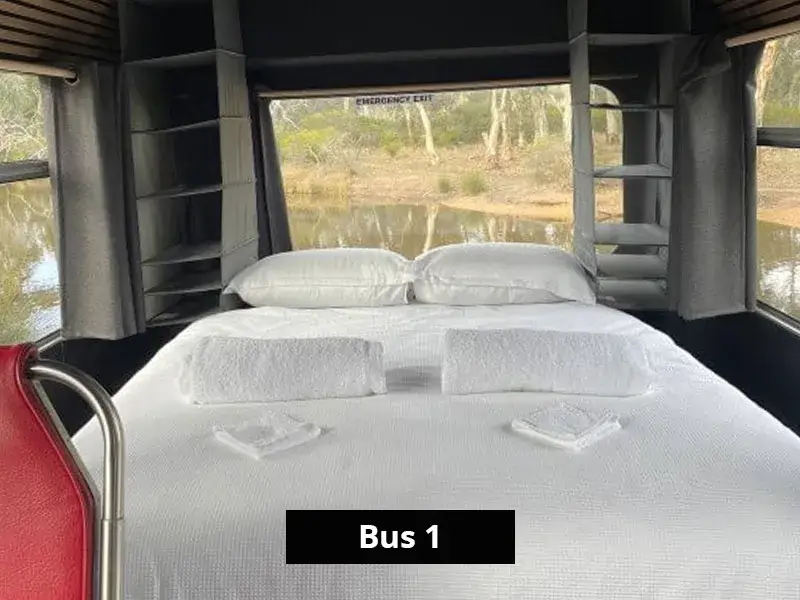 Queen bed inside Bus 1 facing large panoramic windows with lake and bushland views, complete with fresh linen and storage shelves.