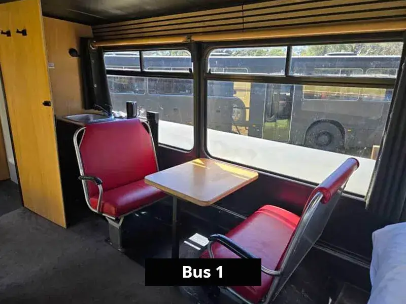 Retro-style dining setup inside Bus 1 with red leather seats, timber table, and large bus windows letting in natural light.