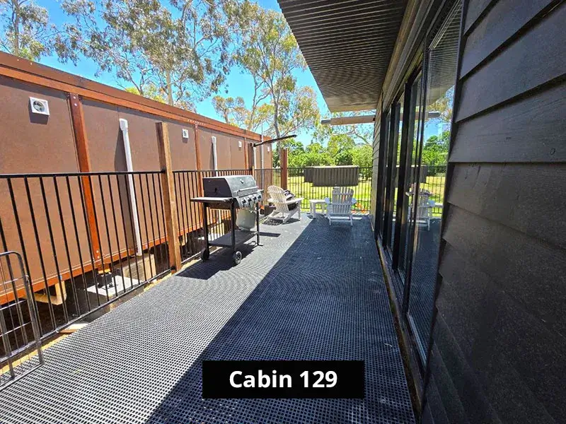 Private deck area of Cabin 129 at Stawell C&C Park, with white Adirondack chairs overlooking a grassy field surrounded by gum trees and nature, enclosed by a black steel fence.