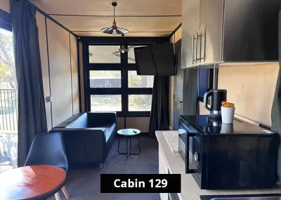 Living area of Cabin 129 at Stawell C&C Park Grampians Gate, featuring a black leather lounge, flat-screen TV mounted on the wall, modern lighting, large rear-facing window, and compact kitchenette with microwave and kettle.