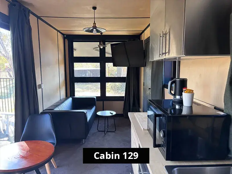 Living area of Cabin 129 at Stawell C&C Park Grampians Gate, featuring a black leather lounge, flat-screen TV mounted on the wall, modern lighting, large rear-facing window, and compact kitchenette with microwave and kettle.