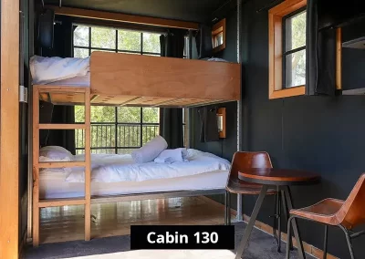 Bunk bed sleeping area in Cabin 130 at Grampians Gate caravan park, featuring wood-framed twin beds with white linens, black interior walls, large windows, and a small round table with two chairs.