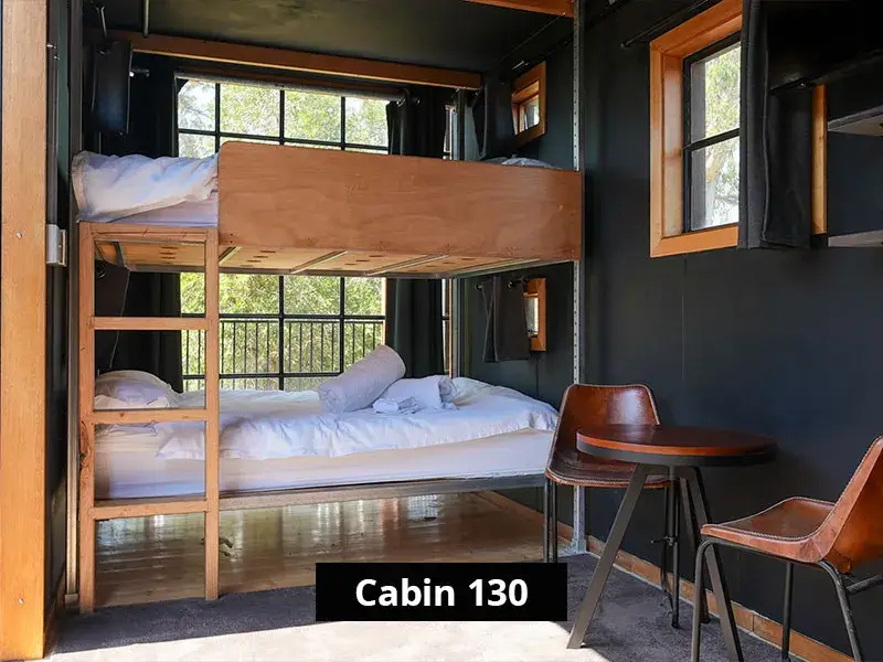 Bunk bed sleeping area in Cabin 130 at Grampians Gate caravan park, featuring wood-framed twin beds with white linens, black interior walls, large windows, and a small round table with two chairs.