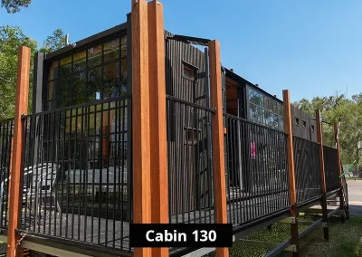 Exterior of Cabin 130 at Stawell C&C Park Grampians Gate, showing a black timber-clad structure elevated on stilts, with vertical wooden posts, metal railings, and large windows facing a tree-lined area.