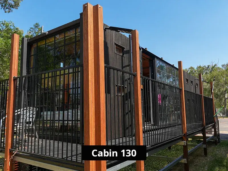 Exterior of Cabin 130 at Stawell C&C Park Grampians Gate, showing a black timber-clad structure elevated on stilts, with vertical wooden posts, metal railings, and large windows facing a tree-lined area.