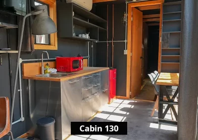 Modern tiny house kitchen with black walls and wood accents, featuring a red microwave, stainless steel cabinets, open shelving with kitchenware, and a small dining area with natural light streaming in – Cabin 130 interior.