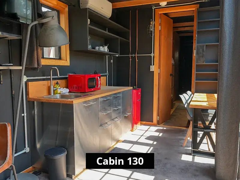 Modern tiny house kitchen with black walls and wood accents, featuring a red microwave, stainless steel cabinets, open shelving with kitchenware, and a small dining area with natural light streaming in – Cabin 130 interior.