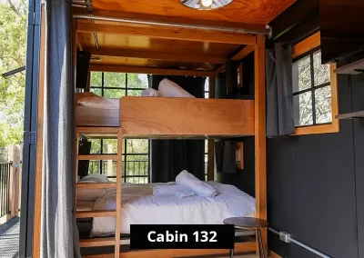 Queen bunk bed setup in Cabin 132 at Stawell C&C Park, featuring timber-framed double and single beds with white linens, surrounded by windows and black interior walls with wood trim – ideal for group or family stays.