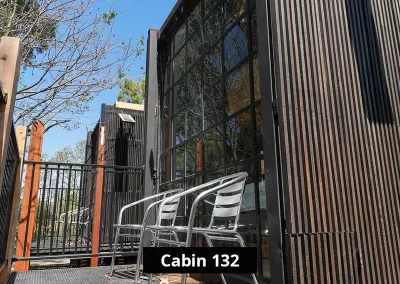 Exterior sitting area of Cabin 132 at Grampians Gate caravan park, showing two metal chairs on a private elevated deck, with black vertical cladding and large industrial-style windows overlooking surrounding trees.