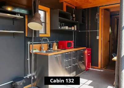 Kitchenette inside Cabin 132 at Stawell C&C Park Grampians Gate, featuring stainless steel drawers, a wood countertop, red microwave and mini fridge, open shelving with glassware and crockery, and industrial-style black walls.
