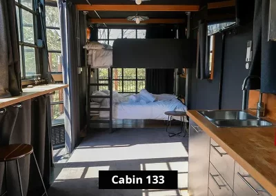 Interior of Cabin 133 at Grampians Gate caravan park showing a compact studio layout with wooden bunk beds, a kitchenette with stainless steel drawers and sink, and large windows allowing in natural light.