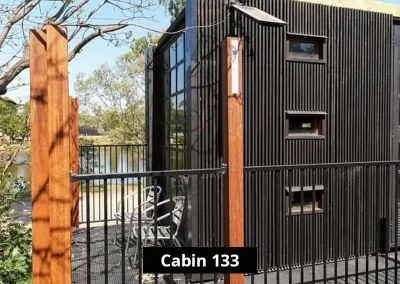Exterior view of Cabin 133 at Stawell C&C Park Grampians Gate, featuring black vertical cladding, narrow windows, and a private elevated deck with two metal chairs overlooking the park’s scenic lakefront.