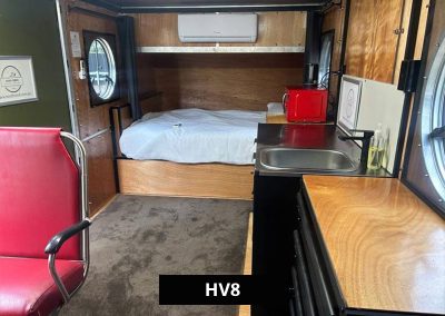 Interior of HV8 tiny home cabin with queen bed, timber walls, compact kitchen with sink, microwave, and round window views.